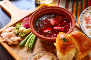 Eine Schale mit Borschtsch steht auf einem Brett auf einem Tisch. Neben der Schale, ebenfalls auf dem Brett liegen Brot, Frühlingszwiebeln, gründe Tomaten und rote Zwiebeln