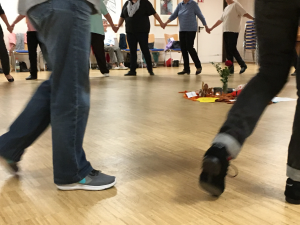 Tanzede Füße im Kreis. Menschen halten sich an den Händen. In der Mitte liegt eine Decke mit Blumen und Kärtchen