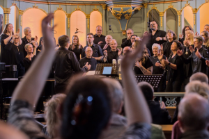 Aus dem Publikum heraus fotografiert mit vordergründiger Unschärfe auf einen Gospelchor, in schwarz gekleidtet in einem Altarraum stehend. Viele klatschende Hände