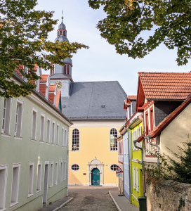 Blick durch eine Gasse auf die Kleine Kirche Alzey. Die Kirche leuchtet im Hintergrund mit ihrer gelben Fassadenfarbe.