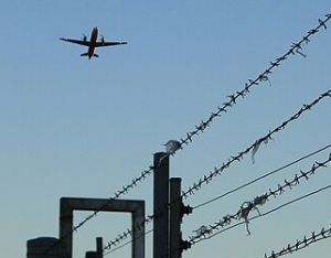 Flugzeug am Himmel, darunter ein Stacheldrahtzaun