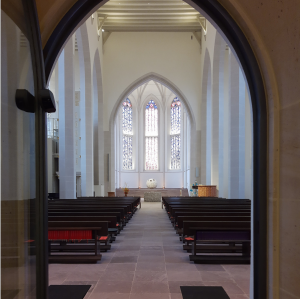 Blick durch eine gläserne Zwischentür in dden Innernraum der Nikolaikirche in Alzey. Man sieht Banreihen und den Altarraum.