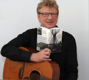 Paddy Schmidt posiert mit seinem Buch in der Hand und der Gitarre vor einer weißen Wand im Halbprofil