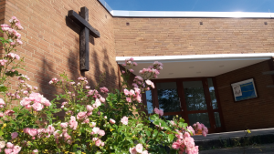 Eingang zum Ev. Gemeindezentrum mit Kreuz an der Wand und blühenden Blumen davor