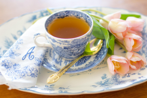 Eine gefüllte Teetasse steht auf einem Kuchenteller, daneben liegt ein goldfarbener Teelöffel.
