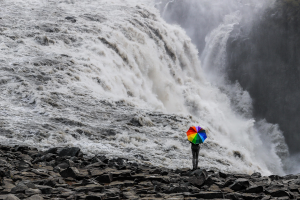 Das Bild zeigt das Schwarzweißfoto eines Wasserfalls. Davor steht ein Mensch mit dem Rücken zur Kamera und einem riesigen Sonnenschirm in Regenbogenfarben.