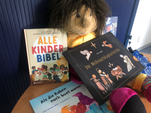 Verschiedene Kinderbücher liegen um eine Handpuppe herum auf einem Tisch. Titel der Bücher: Alle Kinderbibel, Die Weihnachtsgeschichte, Als die Raben noch bunt waren