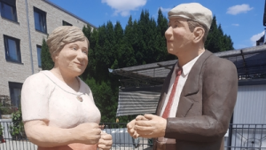 Zwei lebensgroße Skulpturen eines älteren Mannes und einer älteren Frau stehen sich im Freien gegenüber und wirken, als würden sie frei tanzen.